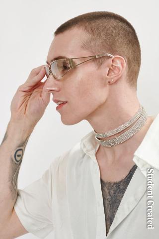 Image of a student photoshoot with a male model with a buzz cut on the side and thicker on on top. They are wearing a sparkling necklace choker and raising their arm to put on sunglassess