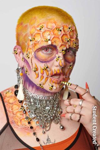 Image of a student photoshoot with a model with full costume makeup of a honey comb on their head with bees on their face. Their hair is style with a buzz cut and colored yellow to an orange ombre to the bottom. Their nails are black and yellow striped with some orange.