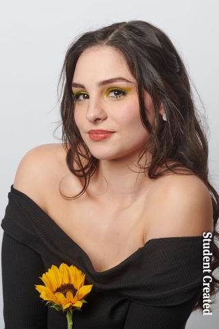 Image of a student photoshoot with a model wearing a black sweater over the shoulder and holding a sunflower. Her hair is styled long and chocolate brown with delicate peachy makeup.