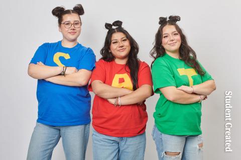 Image of a student photoshoot with three female models. Their hair is done with two buns on top. the model on the left is wearing a blue shirt with an "S". the model in the middle is wearing a shirt with "A" on it and the third model on the right is wearing a green shirt with a "T" on it