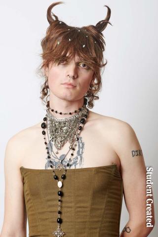 Image of a student photoshoot of a model wearing a brown corset and lots of jewelry. Their hair is light brown with reddish tones and styled short with two horns at the top of their head.