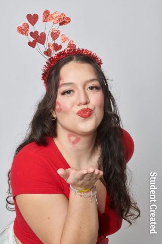 Image of student photoshoot with a female model wearing a red shirt and a headband with hearts. Her hair is long and brown. Her make cheeks have lipstick kisses and she is blowing a kiss